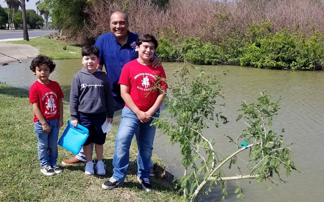 Three young boys stand with their father on the edge of a river beside a young Montezuma cypress tree that has just been planted.