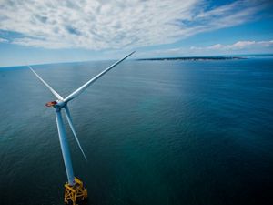 A wind turbine is pictured at left and sits on top of a wide open ocean, with a small patch of land with green grass in view. Clouds are seen on a blue sky.