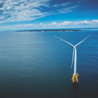 An aerial view of a wind turbine in the ocean with the coast of Block Island and Rhode Island in the background.