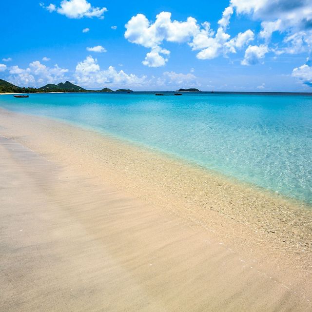 A sandy beach leads to deep blue water, with green mountains on the horizon and boats dotting the shore in the background