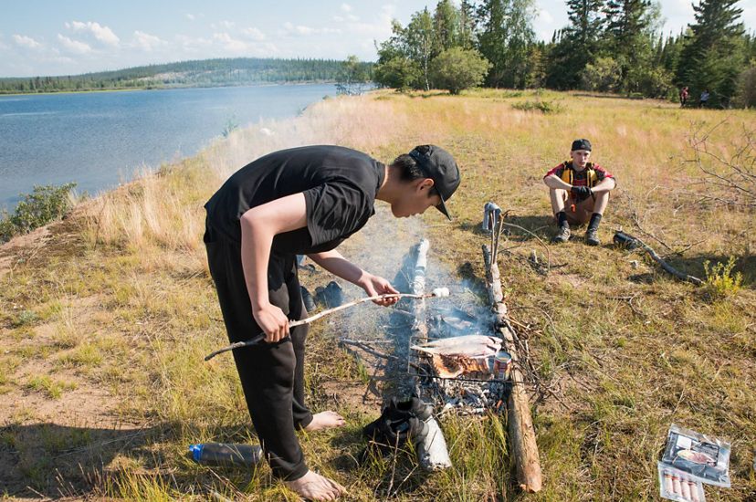 first nation teenage boy roasts marshmallow over fire next to a lake in thaidene nene