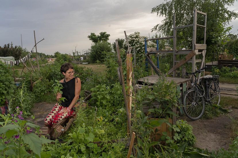 woman wearing black shirt and skirt holds a plant, sits on the ground among many plants with a bicycle nearby