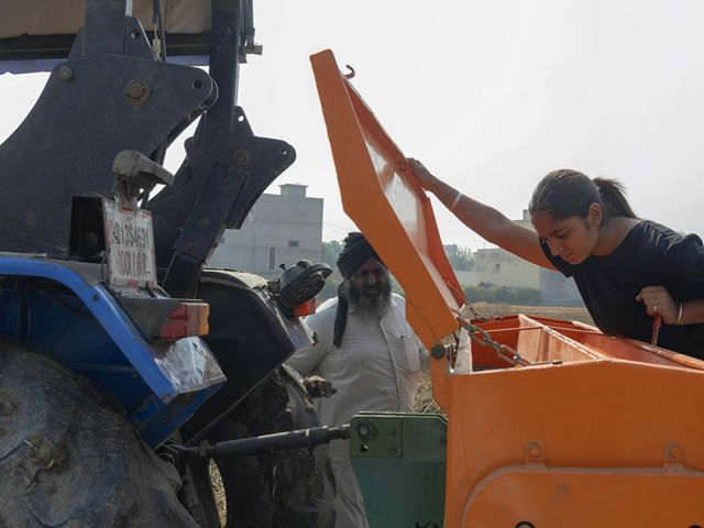 Amandeep lifts a lid from an orange seeder machine and peers inside of the machinery. Her father is nearby.
