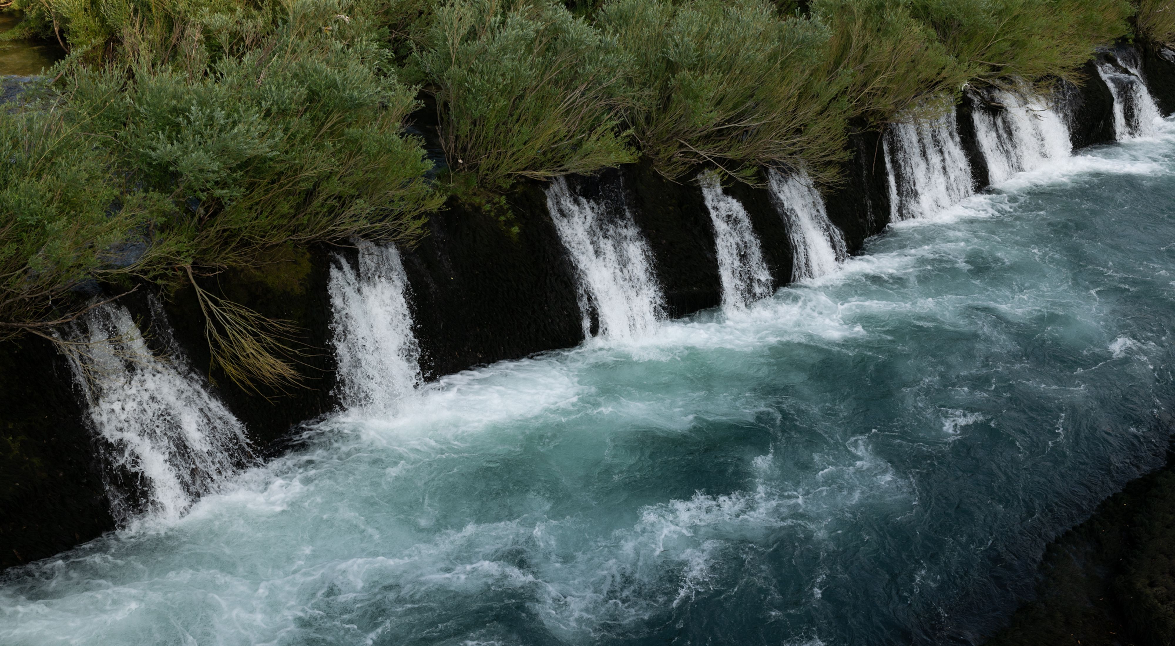 Blue river water cascades and froths over a natural wall of rock.