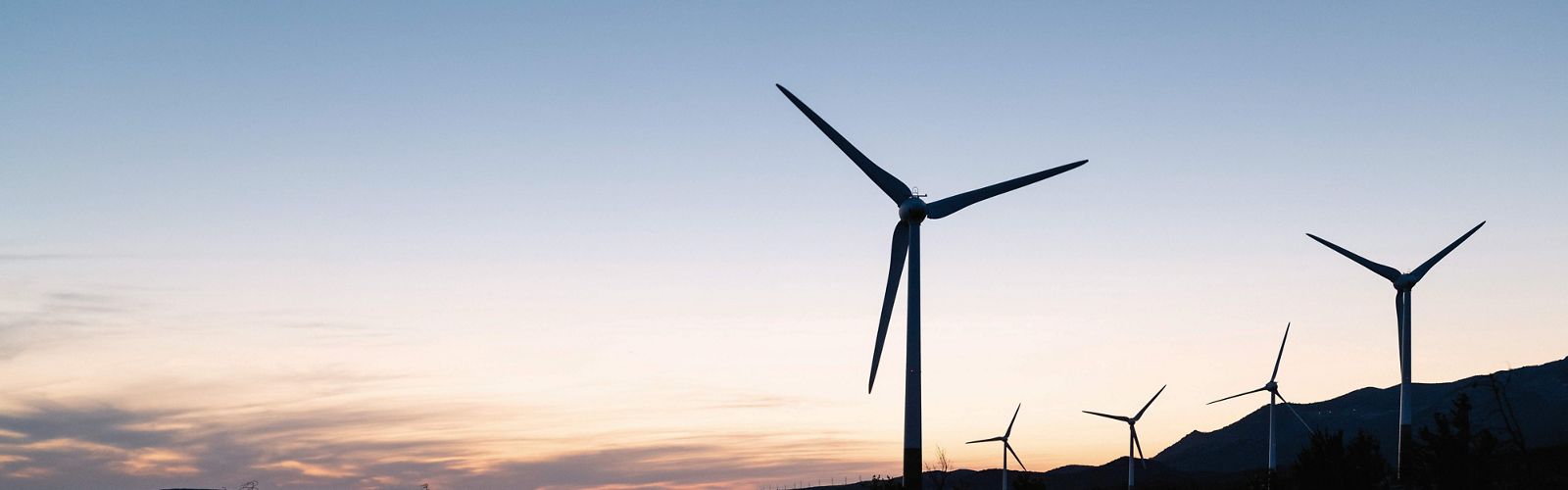 Wind turbines stand on a hilly landscape against an orange sky.